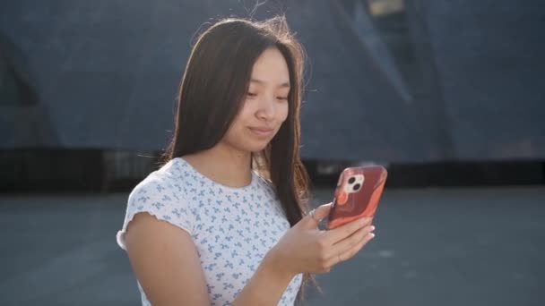 Усміхнена дівчина з Азії користується своїм мобільним телефоном, стоячи надворі.. — стокове відео