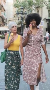 Two mature women enjoying eating an ice-cream while walking together outdoors on the street. Summer concept.