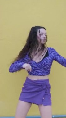 Açık havada sarı duvara karşı hip-hop dansı yaparken kameraya bakan aktif genç bir kadın. Müzik, dans ve gençlik kültürü.