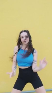 Genç bayan dansçı, dışarıda sarı duvara karşı hip-hop hareketleri yaparken kameraya bakıyor. Müzik, dans ve gençlik kültürü.