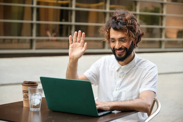 Businessman with makeup smiling and waving hand while having video call on laptop in outdoor cafe. Lgbti, technology and business concept.