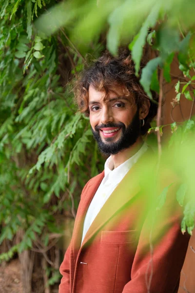 Stylish man with makeup looking at camera and smiling while standing outdoors among green leaves. Lgbti concept.