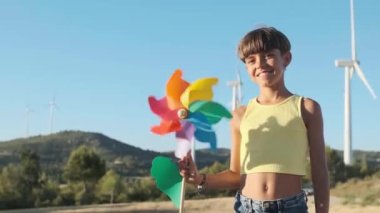 Young girl looking at camera and smiling while holding a wind turbine toy standing outdoors in the field. Renewable energy and sustainable lifestyles concept.
