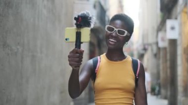 Female influencer with sunglasses streaming on her mobile phone while walking on the street. Technology and social media concept.