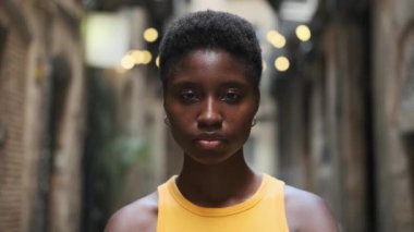 Close-up of serious young woman looking at camera and starting to laugh while standing outdoors.