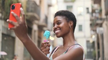 Smiling woman taking selfies with mobile phone while enjoying eating an ice cream on the street. Circular camera movement. Technology and summer concept.