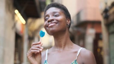 Smiling young woman enjoying eating a lollipop while standing outdoors on the street.