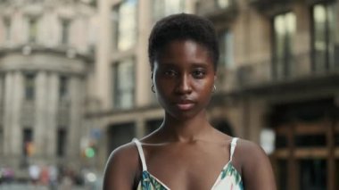 Woman looking at the camera with a serious face and starts to smile as the camera zooms in while standing on the street.