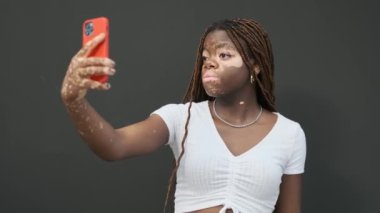 African American woman with vitiligo smiling and showing peace sign while taking selfies with a mobile phone against a dark wall. Technology and social media concept.