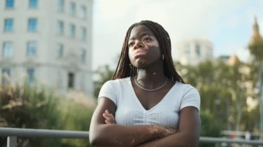 African American woman with vitiligo looking away with a serious expression on her face while standing with her arms crossed outdoors. Circular camera movement.
