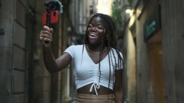 Young female influencer smiling while streaming with her mobile phone walking outdoors on the street. Technology and social networks concept.