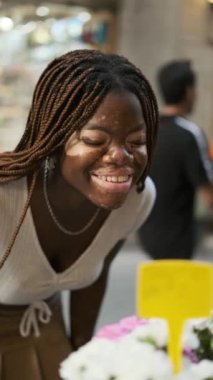 Close up shot of a young African American woman with vitiligo smiling while smelling flowers outdoors at a flower shop.