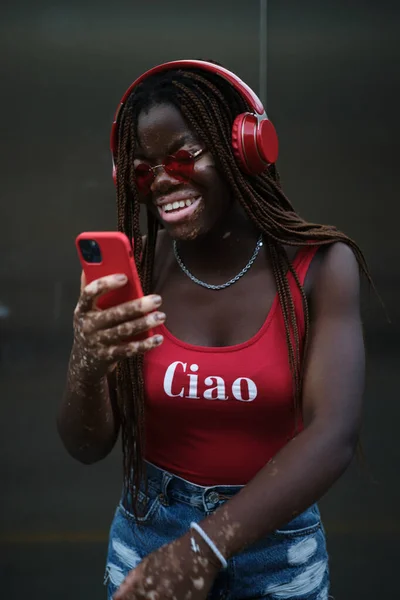 African American woman with vitiligo wearing sunglasses and smiling while using a mobile phone with wireless headphones. Technology concept.