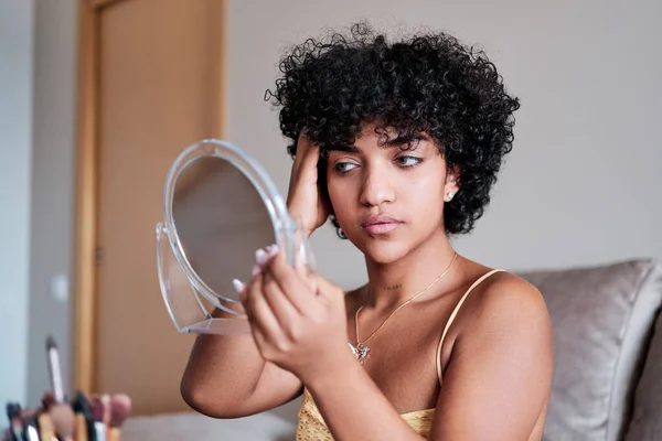 Transgender woman looking in a small mirror and combing her hair while getting ready at home.