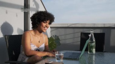 Terasta oturan bir dizüstü bilgisayarla video görüşmesi yapan transseksüel bir kadın el sallıyor ve gülümsüyor. Teknoloji ve iş konsepti.