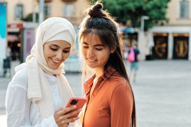 İki genç, çok ırklı bayan arkadaş sokakta yan yana cep telefonu kullanırken gülümsüyor. Teknoloji, arkadaşlık ve sosyal medya kavramı.