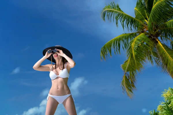 Beautiful young woman in bikini looking through binoculars at the tropical beach — Stock Photo, Image