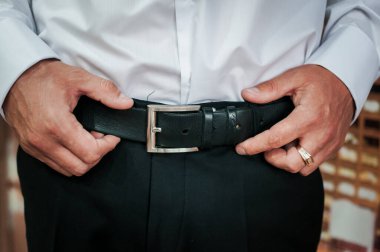 Damat kemerini sıkıyor. Ellerini kemerinde tutan bir adam.