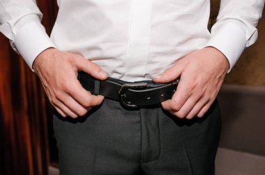 Damat kemerini sıkıyor. Ellerini kemerinde tutan bir adam.