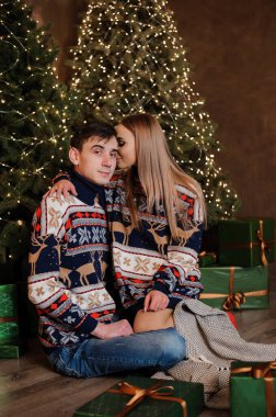 Bir erkek ve bir kız, bir Noel ağacı ve kırmızı bir kanepenin arka planında sarılıp öpüşürler.