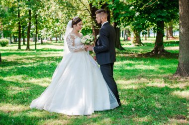Gelin ve damat yazın parkta yürüyorlar, sarılıyorlar. Yeni evliler parkta yürüyorlar da. Arka planda ağaçlar