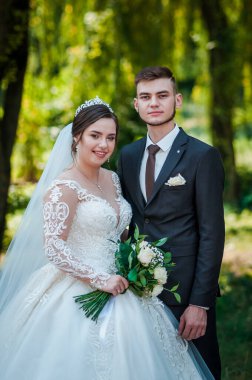 Gelin ve damat yazın parkta yürüyorlar, sarılıyorlar. Yeni evliler parkta yürüyorlar da. Arka planda ağaçlar