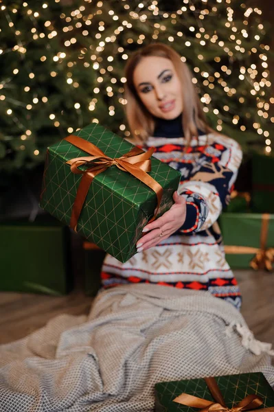 Dziewczyna w swetrze z jeleniem siedzi na podłodze i trzyma zielone pudełko z prezentami. Choinki w tle — Zdjęcie stockowe