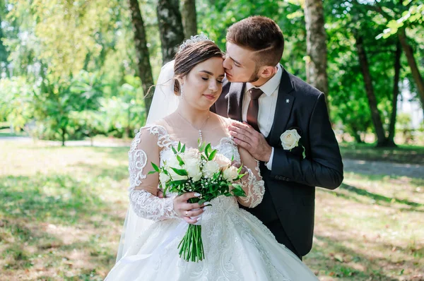 Das Brautpaar spaziert im Sommer im Park und umarmt sich. Die Frischvermählten gehen im Park spazieren. Bäume im Hintergrund — Stockfoto