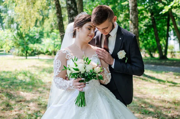 Nevěsta a ženich se v létě procházejí v parku a objímají se. Novomanželé se procházejí v parku. Stromy v pozadí — Stock fotografie