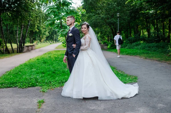 Les mariés se promènent dans le parc en été, en s'embrassant. Les jeunes mariés marchent dans le parc. Arbres en arrière-plan — Photo