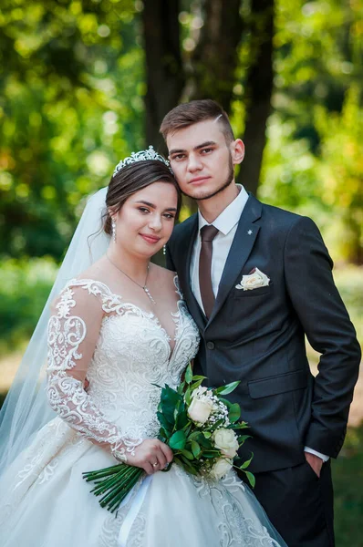 Gelin ve damat yazın parkta yürüyorlar, sarılıyorlar. Yeni evliler parkta yürüyorlar da. Arka planda ağaçlar — Stok fotoğraf