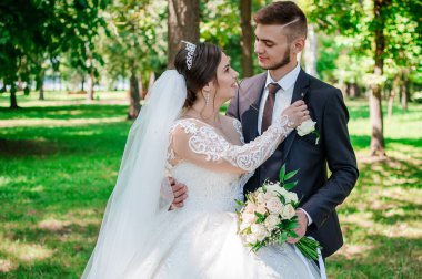 Gelin ve damat yazın parkta yürüyorlar, sarılıyorlar. Yeni evliler parkta yürüyorlar da. Arka planda ağaçlar