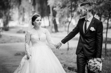 Gelin ve damat yazın parkta yürüyorlar, sarılıyorlar. Yeni evliler parkta yürüyorlar da. Arka planda ağaçlar