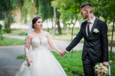 Gelin ve damat yazın parkta yürüyorlar, sarılıyorlar. Yeni evliler parkta yürüyorlar da. Arka planda ağaçlar