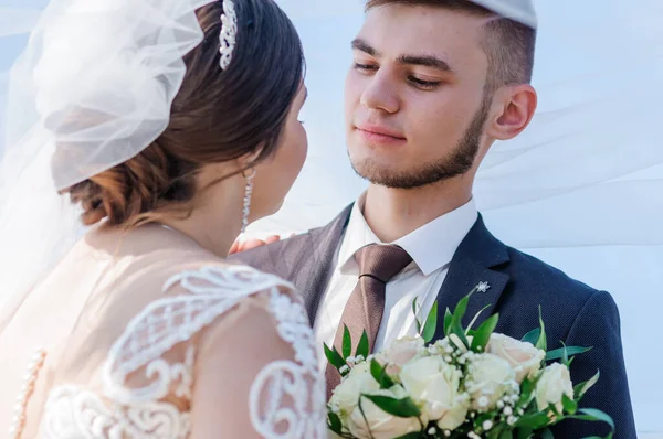 Novomanželé se objímají a líbají pod závojem. Nevěsta drží kytici květin — Stock fotografie