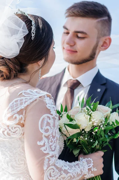 De pasgetrouwden knuffelen en kussen onder de sluier. Bruid met een boeket bloemen — Stockfoto