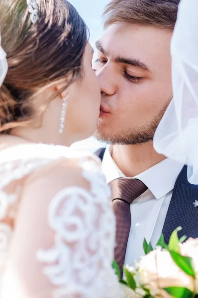 Novomanželé se objímají a líbají pod závojem. Nevěsta drží kytici květin — Stock fotografie