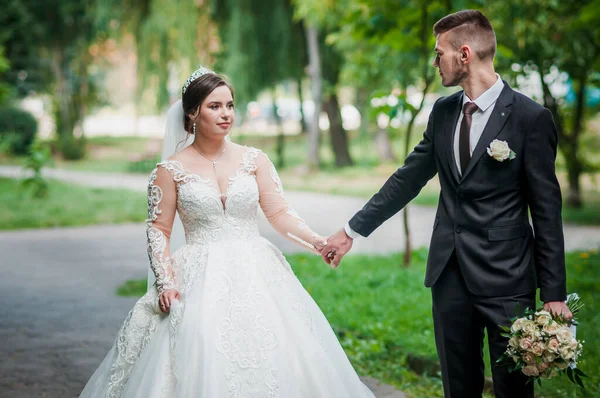 Gelin ve damat yazın parkta yürüyorlar, sarılıyorlar. Yeni evliler parkta yürüyorlar da. Arka planda ağaçlar — Stok fotoğraf