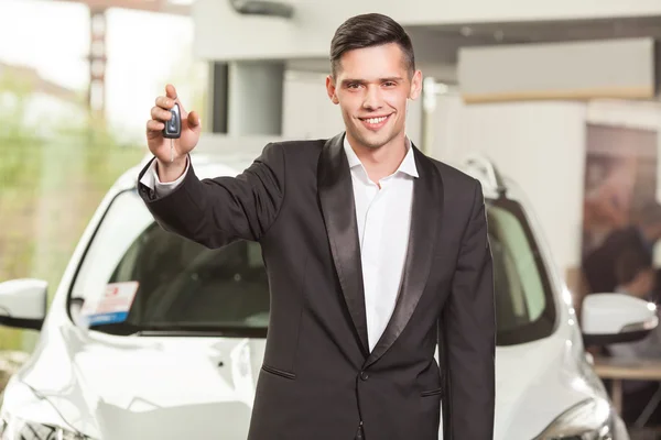 Grande escolha! Bonito jovem vendedor de carros clássico em pé na concessionária segurando uma chave — Fotografia de Stock