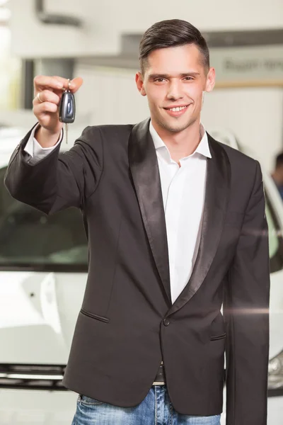 Grande escolha! Bonito jovem vendedor de carros clássico em pé na concessionária segurando uma chave — Fotografia de Stock