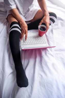 Siyah diz boyu çorap giyen bir kadın yatağında oturup defter bilgisayarı üzerinde çalışıyor.