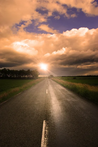 ถนนยางมะตอยที่มีเมฆและรังสีของแสงแดดในพระอาทิตย์ตก — ภาพถ่ายสต็อก