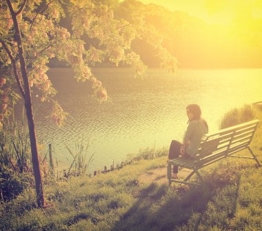 Woman with lake in front of her while sitting on a park bench clipart