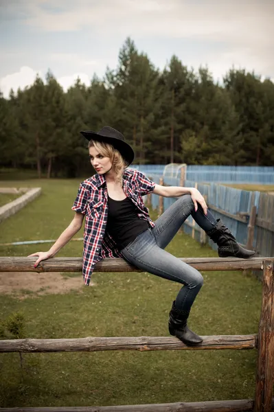 Young girl wearing jeans and checkered shirt sitting on the fence — Stock Photo, Image