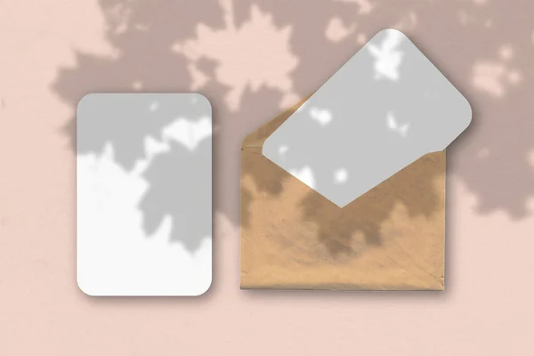 Ein Umschlag mit zwei Blättern strukturierten weißen Papiers auf rosa Tischhintergrund. Mockup mit einer Überlagerung von Pflanzenschatten. Natürliches Licht wirft Schatten einer tropischen Pflanze. — Stockfoto