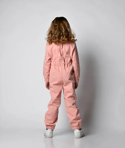 Vista trasera Foto de perfil de longitud completa de una chica rubia rizada positiva de pie en zapatillas blancas y un mono de algodón rosa sobre un fondo claro Imagen de archivo