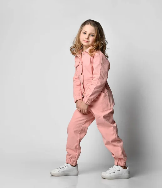 Volledige profielfoto van een positief krullend blond meisje in witte sneakers en roze katoenen overalls, halve zijde op een lichte achtergrond — Stockfoto