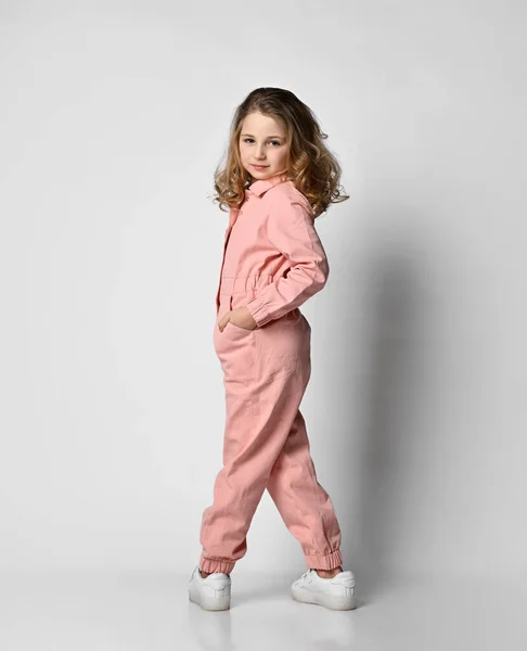 Meisje in een roze katoenen jumpsuit staat met haar rug naar de camera en draaide met haar benen wijd uit elkaar, haar hand in haar zak. — Stockfoto