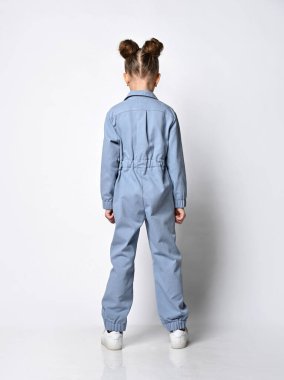 Beyaz spor ayakkabılı ve mavi pamuklu bir tulumla arka planda duran pozitif bir kızın tam boy profil fotoğrafı.