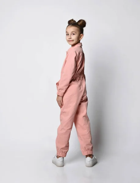 Meisje in een roze katoenen jumpsuit staat met haar rug naar de camera en draaide met haar benen wijd uit elkaar tegen een lichte achtergrond — Stockfoto